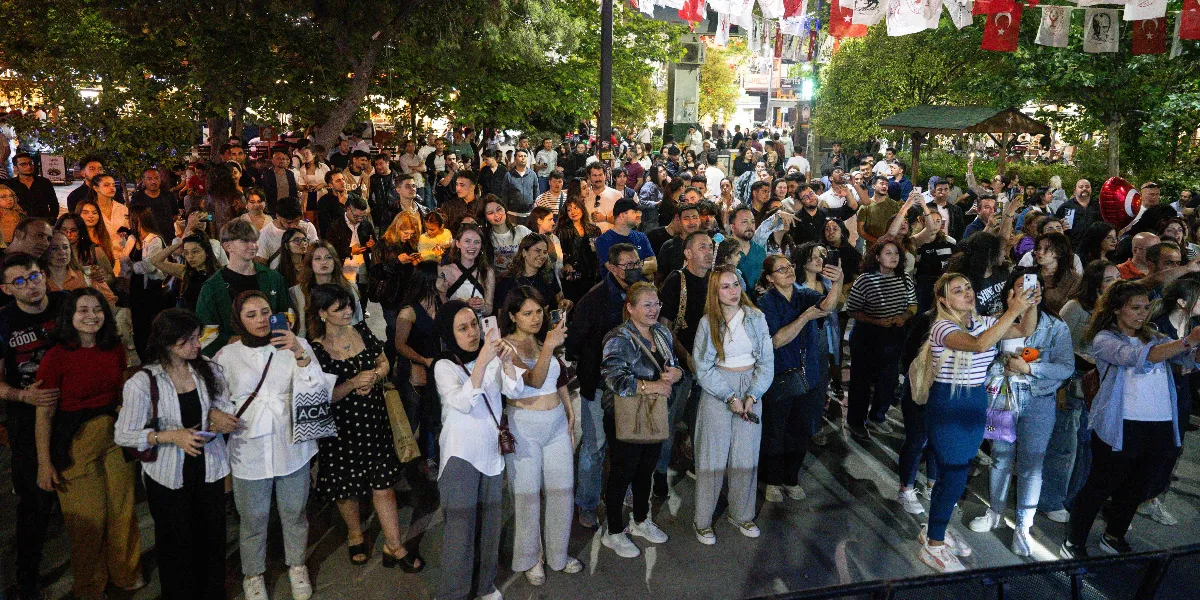 Akşam saatlerinde açık havada düzenlenen bir etkinlikte, sahneyi izleyen ve cep telefonlarıyla görüntü kaydeden kalabalık bir genç grubu. İnsanların çoğu ayakta duruyor, birçoğu ellerindeki telefonlarla çekim yapıyor veya sahneye odaklanmış durumda. Arka planda ağaçlar ve yukarıdan sarkan Türk bayrakları ile süslü bir alan görülüyor.
