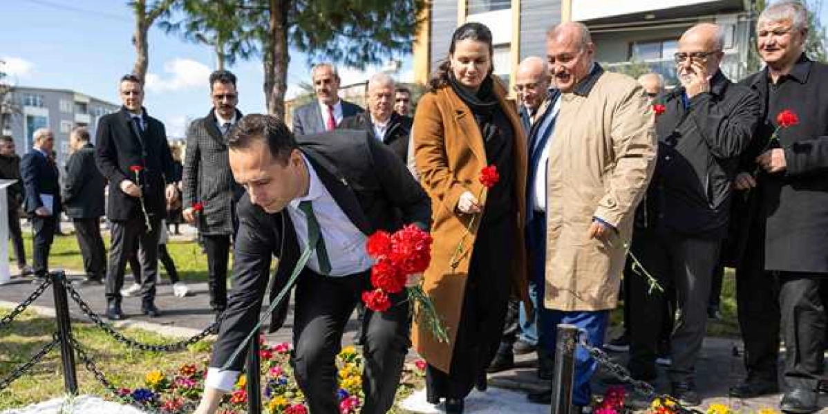 Açık havada, bir anıtın önünde düzenlenen anma töreninde, takım elbiseli bir erkek elindeki kırmızı karanfilleri yere bırakıyor. Yanında ve arkasında, ellerinde kırmızı karanfiller tutan, resmi giyimli bir grup kadın ve erkek, saygı duruşu içerisinde anıtı izliyor. Arka planda ağaçlar ve modern binalar görülüyor.