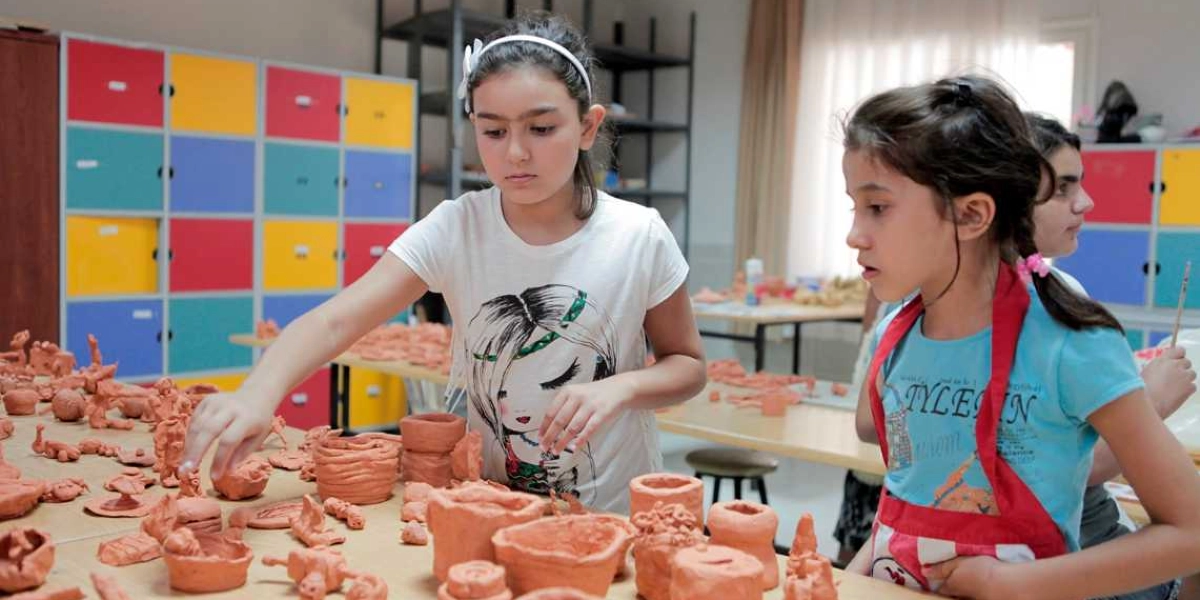 Sanat atölyesinde kil hamuruyla çeşitli figürler ve kaplar yapan çocuklar, masanın üzerinde duran çalışmalarını dikkatle inceliyor. Arka planda renkli bölmeli dolaplar bulunan sınıfta, beyaz tişörtlü bir kız çocuğu elindeki kil parçasını şekillendirirken, yanında kırmızı önlüklü mavi tişörtlü bir arkadaşı ona eşlik ediyor.