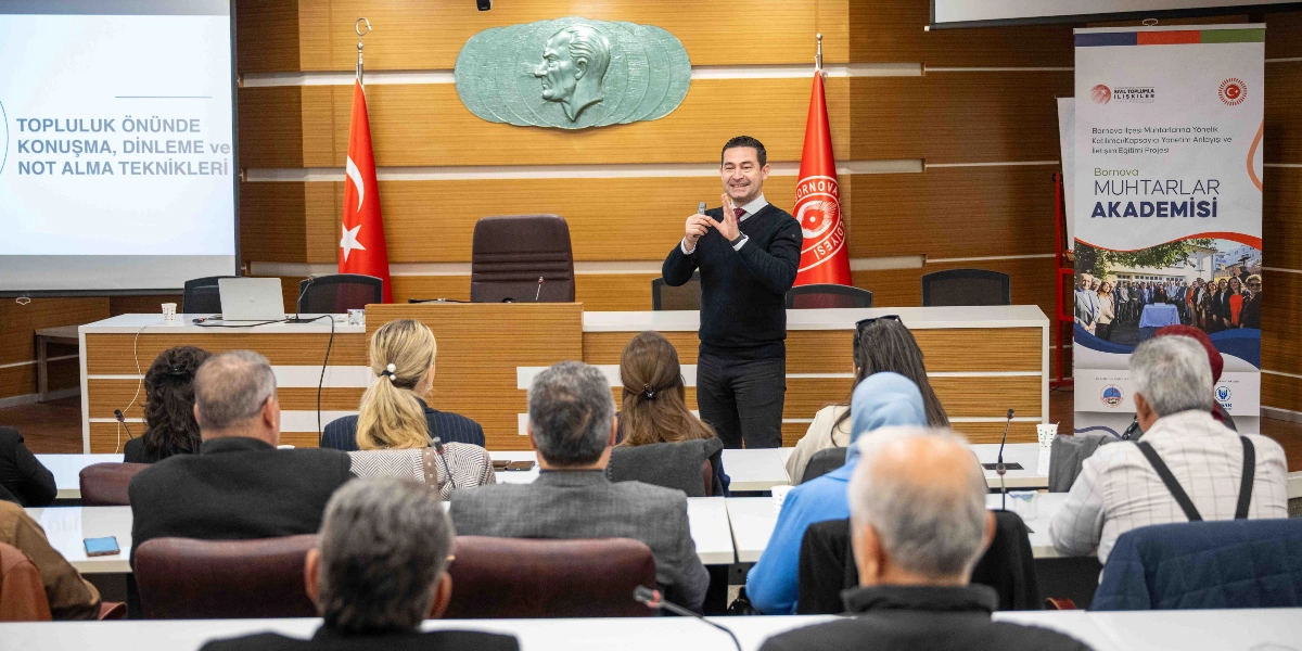 Bornova Muhtarlar Akademisi kapsamında düzenlenen bir eğitim toplantısında, kürsüde duran bir konuşmacı katılımcılara hitap ediyor. Konuşmacının arkasındaki duvarda Atatürk kabartması, iki yanında Türk bayrağı ve Bornova Belediyesi flaması yer alıyor. Soldaki projeksiyon ekranında 