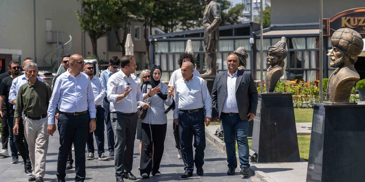 Bornova'da bir parkta yürüyen ve sohbet eden bir grup insan. Grubun önünde beyaz gömlekli bir erkek, yanında başörtülü bir kadın ve diğer katılımcılar yer alıyor. Arka planda heykellerin bulunduğu bir yürüyüş yolu ve bir restoran binası görülüyor.