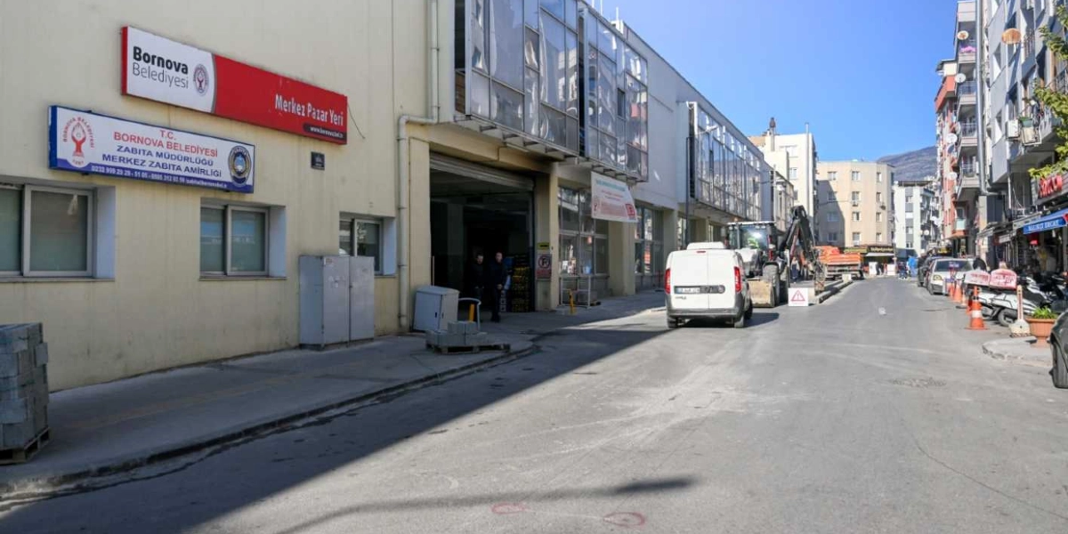 Bornova Belediyesi Merkez Pazar Yeri ve Zabıta Amirliği binasının önünde devam eden yol düzenleme çalışmaları. Sokak üzerinde beyaz bir hafif ticari araç, bir iş makinesi ve yol kenarında trafik konileri bulunuyor. Binanın dış cephesinde belediyenin tabelaları yer alıyor, arka planda ise çok katlı konutlar görünüyor.