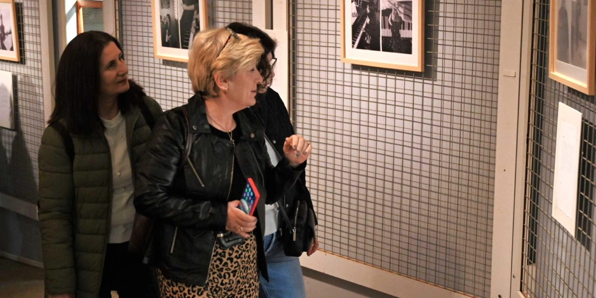 Bir sanat galerisinde, ızgara paneller üzerine asılmış siyah beyaz fotoğrafları inceleyen üç kadın. Önde duran sarı saçlı kadın, elinde bir akıllı telefon tutarak yanındaki arkadaşına duvardaki bir eseri gösteriyor. Yanındaki koyu renk ceketli kadınlar ise ilgiyle fotoğraflara bakıyor.