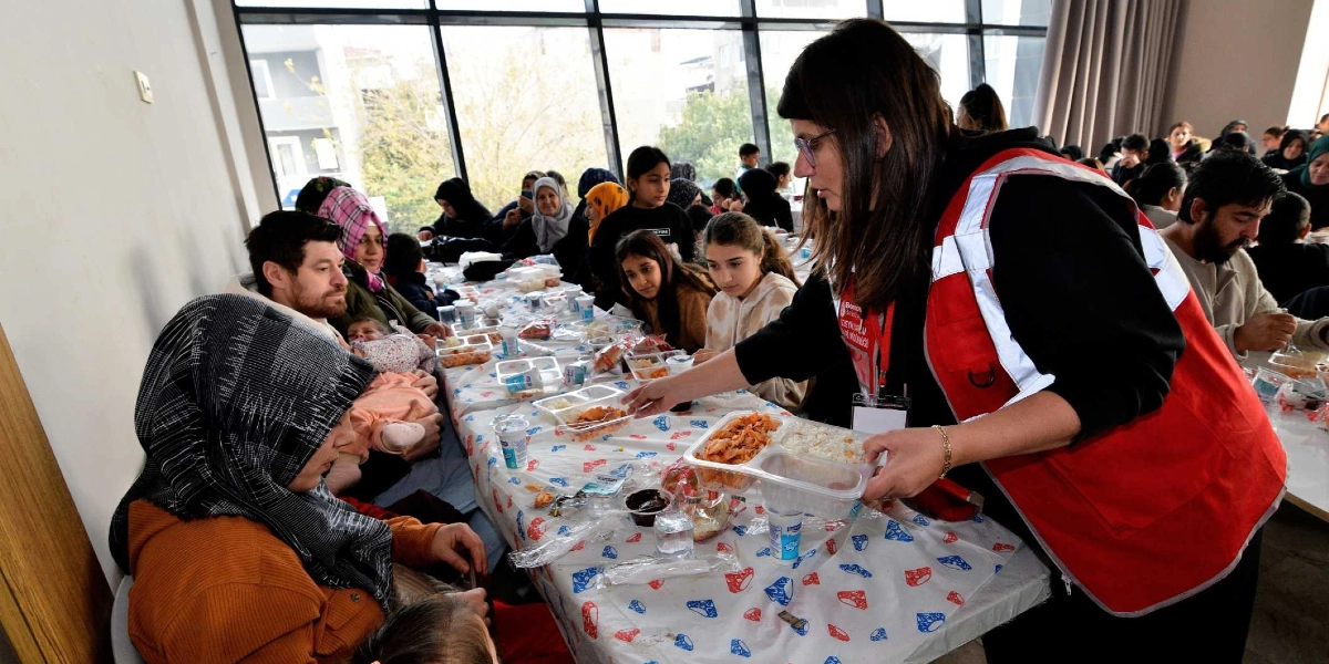 Bornova Belediyesi logolu kırmızı yelek giyen bir görevli, uzun bir masada oturan vatandaşlara plastik kaplarda yemek servisi yapıyor. Masada başörtülü kadınlar, çocuklar ve bebekli aileler oturuyor; önlerinde ise yemek kapları ve içecekler bulunuyor. Arka planda boydan boya camlı bir pencere ve içeride kalabalık bir yemek ortamı görülüyor.
