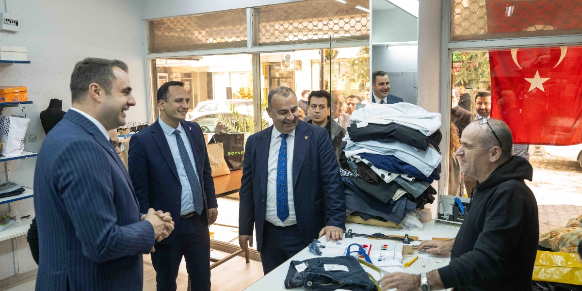 Takım elbiseli üç CHP'li siyasetçi, bir terzi dükkanında çalışan bir esnafla sohbet ediyor. Siyasetçiler gülümseyerek esnafı dinlerken, esnaf tezgahın üzerindeki kıyafetlerin yanında duruyor. Dükkanın camında asılı bir Türk bayrağı bulunuyor ve arka planda dışarıdaki cadde görünüyor.