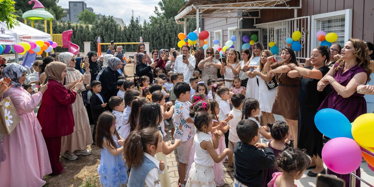 Bir anaokulunun bahçesinde düzenlenen karne töreninde, çocuklar ve öğretmenleri müzik eşliğinde dans ediyor. Renkli balonlarla süslenmiş alanda, öğretmenler ve veliler çocukların dansını izlerken cep telefonlarıyla bu anları kaydediyor. Ön planda neşeyle hareket eden çocuklar, arka planda ise onları izleyen yetişkinler yer alıyor.