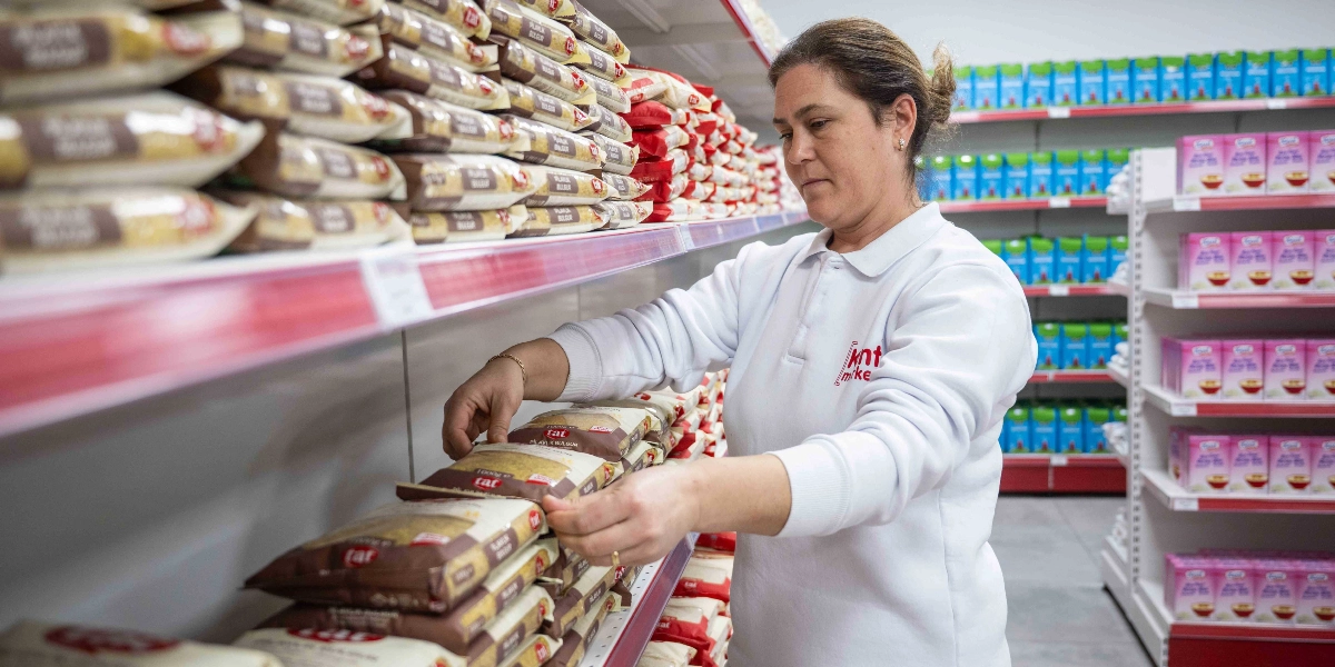 Üzerinde Kent Market logosu bulunan beyaz bir iş kıyafeti giyen kadın çalışan, market rafındaki Tat marka bulgur paketlerini düzenliyor. Raflar, benzer paketlenmiş gıda ürünleriyle dolu.