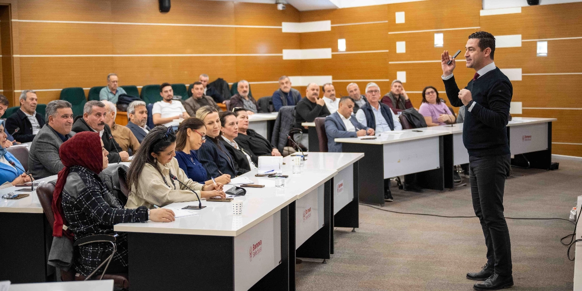 Modern bir konferans salonunda, Bornova Belediyesi logosunun yer aldığı masalarda oturan bir grup muhtar, ayakta sunum yapan bir konuşmacıyı dinliyor. Konuşmacı, koyu renkli kazağı ve gömleğiyle sağ elini havaya kaldırarak bir noktaya işaret ediyor. Katılımcılar arasında kadın ve erkek muhtarlar bulunuyor; bazıları not alıyor, bazıları ise dikkatle konuşmacıya bakıyor. Salonun duvarları ahşap panellerle kaplı ve ortamda resmi bir eğitim veya toplantı atmosferi hakim.