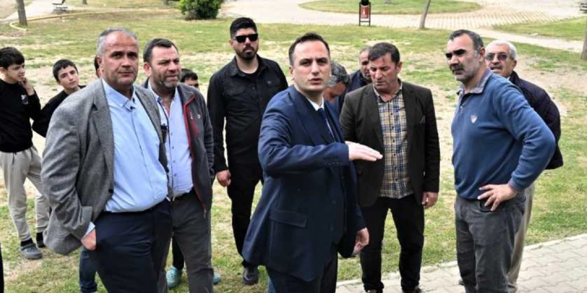 Bornova Belediye Başkanı Ömer Eşki, bir park alanında yanındaki heyetle birlikte incelemelerde bulunuyor. Lacivert takım elbiseli Eşki, eliyle çevreyi işaret ederek konuşurken, çevresinde farklı kıyafetler giymiş bir grup erkek ve arka planda gençler yer alıyor.
