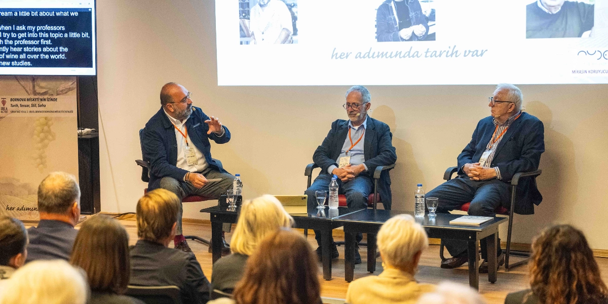 Bir panelde konuşan üç erkek katılımcı, sahnede yan yana dizilmiş sandalyelerde oturuyor. Soldaki katılımcı el hareketleriyle bir şeyler anlatırken, ortadaki ve sağdaki katılımcılar onu dinliyor. Katılımcıların önünde üzerinde su bardakları ve bir tablet bulunan siyah bir masa yer alıyor. Arka planda, üzerinde 