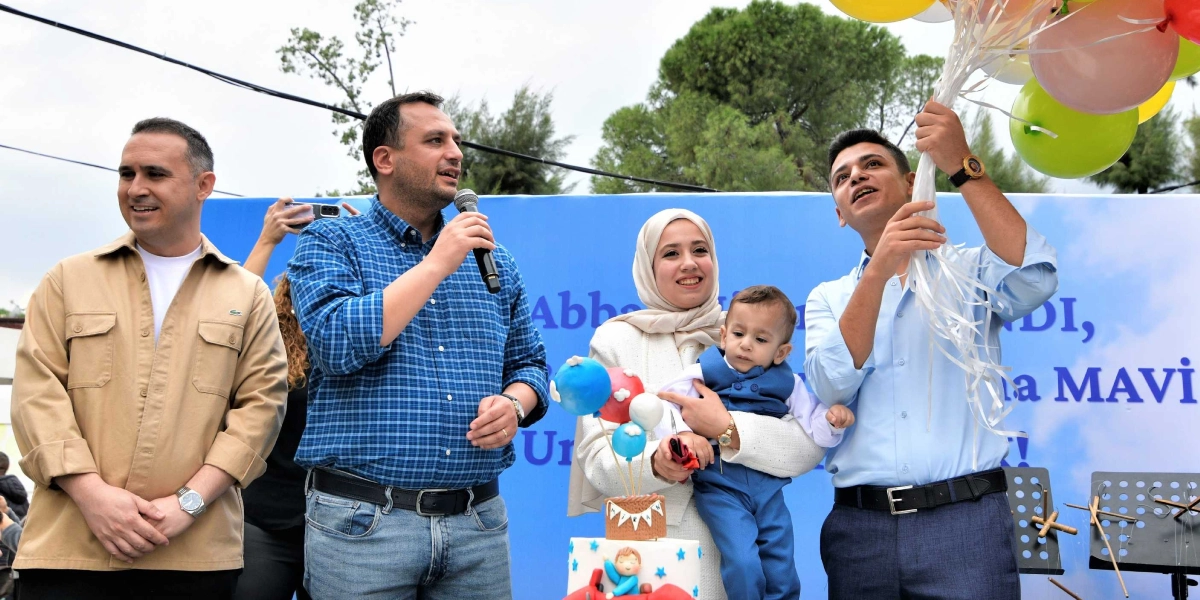 Mavi bir fon önünde duran dört kişilik bir grup; solda bej ceketli bir adam, yanında mikrofonla konuşan kareli gömlekli bir adam, ortada kucağında bebek tutan başörtülü bir kadın ve sağda gökyüzüne doğru renkli balonlar bırakan açık mavi gömlekli bir genç adam yer alıyor. Kadının önünde üzerinde küçük balon süslemeleri ve 