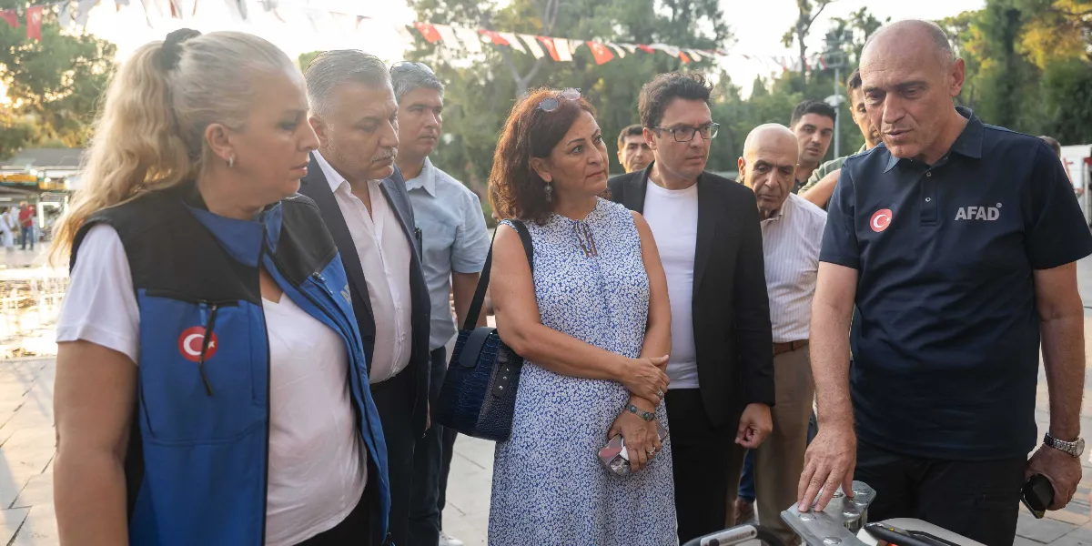 Açık havada, üzerinde AFAD logolu lacivert bir tişört bulunan bir yetkili, yanındaki gruba teknik bir ekipmanı göstererek bilgi veriyor. Grubun önünde mavi yelekli bir kadın, takım elbiseli bir erkek ve desenli bir elbise giyen bir kadın dikkatle dinliyor; arka planda ise diğer katılımcılar ve Türk bayraklarıyla süslenmiş bir alan yer alıyor.