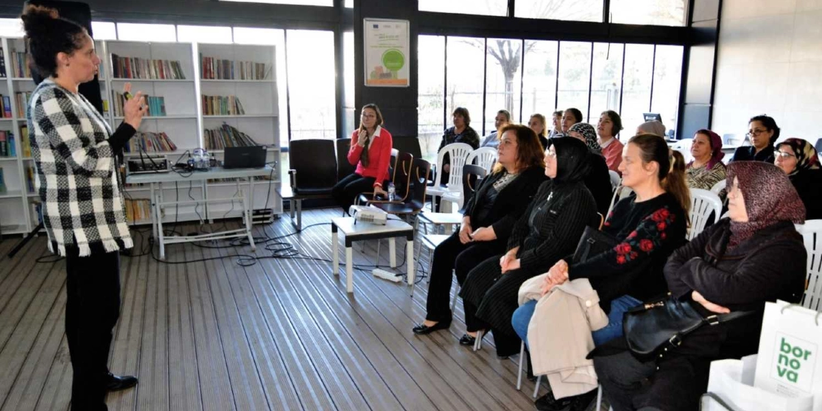 Aydınlık ve geniş bir kütüphane ortamında, kareli hırka giymiş bir kadın ayakta durarak katılımcılara sunum yapıyor. Karşısında beyaz sandalyelerde oturan, farklı yaş gruplarından kadınlardan oluşan bir grup, dikkatle konuşmacıyı dinliyor. Arka planda kitaplarla dolu raflar, büyük pencereler ve bir projeksiyon cihazı yer alıyor. Ön planda ise üzerinde 
