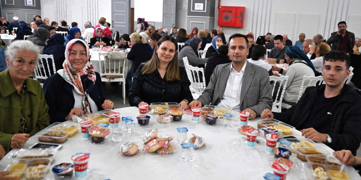Geniş bir salonda düzenlenen dayanışma yemeğinde, beyaz örtülü yuvarlak bir masanın etrafında oturan beş kişilik bir grup kameraya bakıyor. Masanın üzerinde plastik kaplarda servis edilmiş yemekler, Çeker markalı yoğurtlar ve ekmekler bulunuyor. Arka planda ise benzer şekilde masalarda oturan çok sayıda insan yer alıyor.