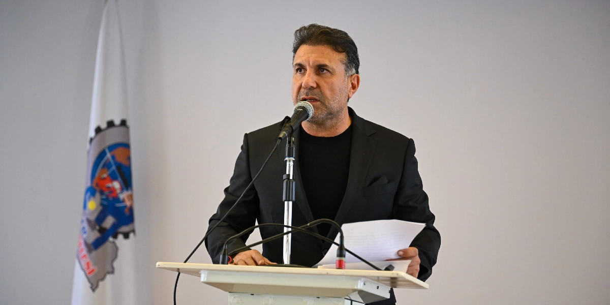 Siyah ceket ve siyah tişört giymiş orta yaşlı bir erkek, kürsüde mikrofon başında konuşma yapıyor. Elinde bir kağıt tutan konuşmacının sol tarafında, üzerinde çark ve dünya figürü bulunan beyaz bir sanayi sitesi bayrağı yer alıyor.