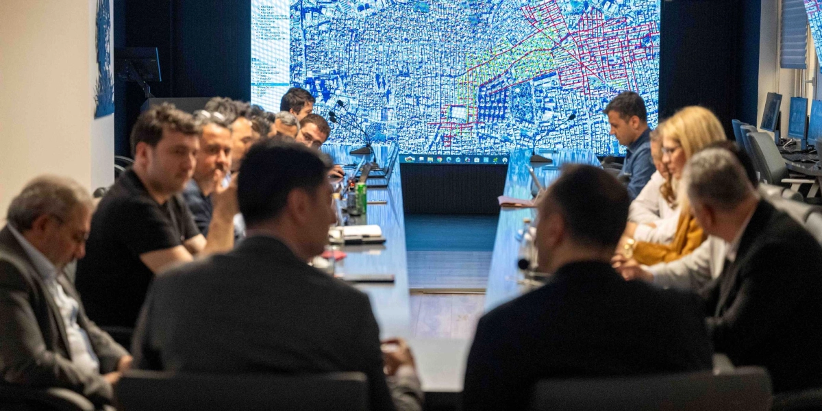 Modern bir toplantı odasında, uzun bir masa etrafında toplanmış bir grup insan, karşı duvardaki dev ekranda yer alan detaylı bir şehir haritası ve dijital ikiz verilerini inceleyerek toplantı yapıyor.