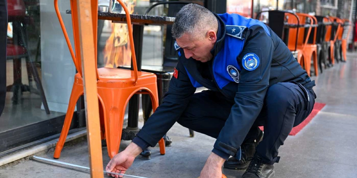 Bornova Belediyesi zabıta üniformalı bir görevli, bir kafe önündeki kaldırımda şerit metre kullanarak ölçüm yapıyor. Görevli, turuncu metal sandalyelerin yanındaki alanda denetim gerçekleştiriyor.