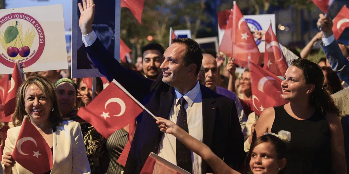 Bornova Belediye Başkanı Ömer Eşki, kalabalık bir kortej yürüyüşünde el sallayarak halkı selamlıyor. Çevresindeki insanlar ellerinde Türk bayrakları ve Atatürk posterleri taşıyor. Sol tarafta üzerinde kiraz görseli bulunan 
