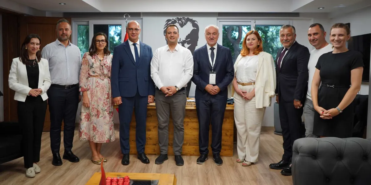 Ofis ortamında yan yana duran, aralarında Bornova Belediyesi yetkilileri ve Bosna-Hersek heyetinin bulunduğu dokuz kişilik grup, arka planda Atatürk silüetinin yer aldığı bir masanın önünde poz veriyor. Gruptaki erkekler takım elbise veya gömlek, kadınlar ise şık elbiseler ve ceketler giymiş durumda.