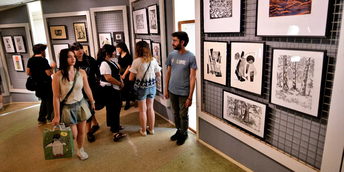 Bornova'da düzenlenen İnce İşler sergisinde, duvarlardaki metal ızgaralara asılmış siyah beyaz baskı eserleri inceleyen bir grup genç ziyaretçi yer alıyor. Ön planda elinde üzerinde penguen çizimi olan bir alışveriş çantası taşıyan genç bir kadın, arka planda ise sergilenen sanat eserlerine bakan diğer ziyaretçiler görülüyor.