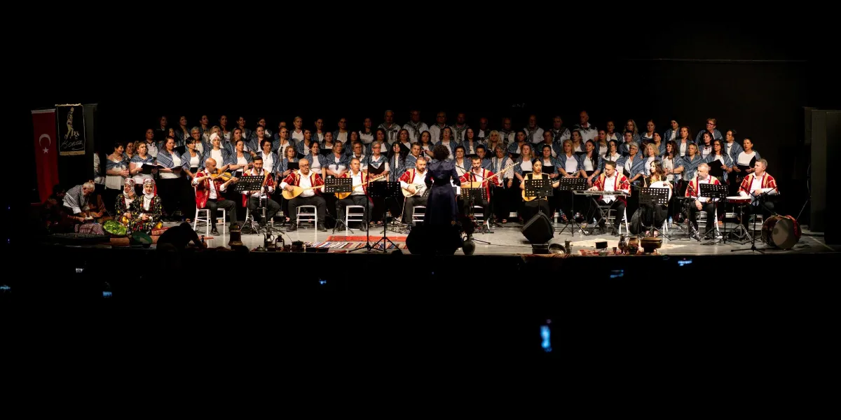 Karanlık bir sahnede, geleneksel kıyafetler giymiş geniş bir koro ve orkestra grubu performans sergiliyor. Önde, saz, keman ve çeşitli enstrümanlar çalan müzisyenler, arkada ise ayakta duran koro üyeleri yer alıyor. Sahnenin ortasında orkestra şefi sırtı dönük bir şekilde yönetimi sağlıyor. Sol tarafta Türk bayrağı ve üzerinde Bornova yazılı bir flama bulunuyor. Sahne zemini, kilimler ve bazı geleneksel objelerle dekore edilmiş.