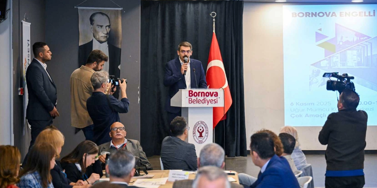 Bornova Belediyesi logolu kürsüde konuşma yapan gözlüklü bir erkek, arkasında Türk bayrağı ve Mustafa Kemal Atatürk portresi ile izleyicilere hitap ediyor. Salonun sağ tarafındaki yansıtıcı ekranda 