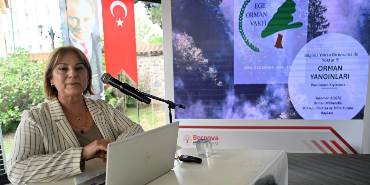 Açık renkli çizgili ceket giymiş bir kadın, önündeki dizüstü bilgisayarla bir sunum yaparken mikrofon başında oturuyor. Arka planda üzerinde 