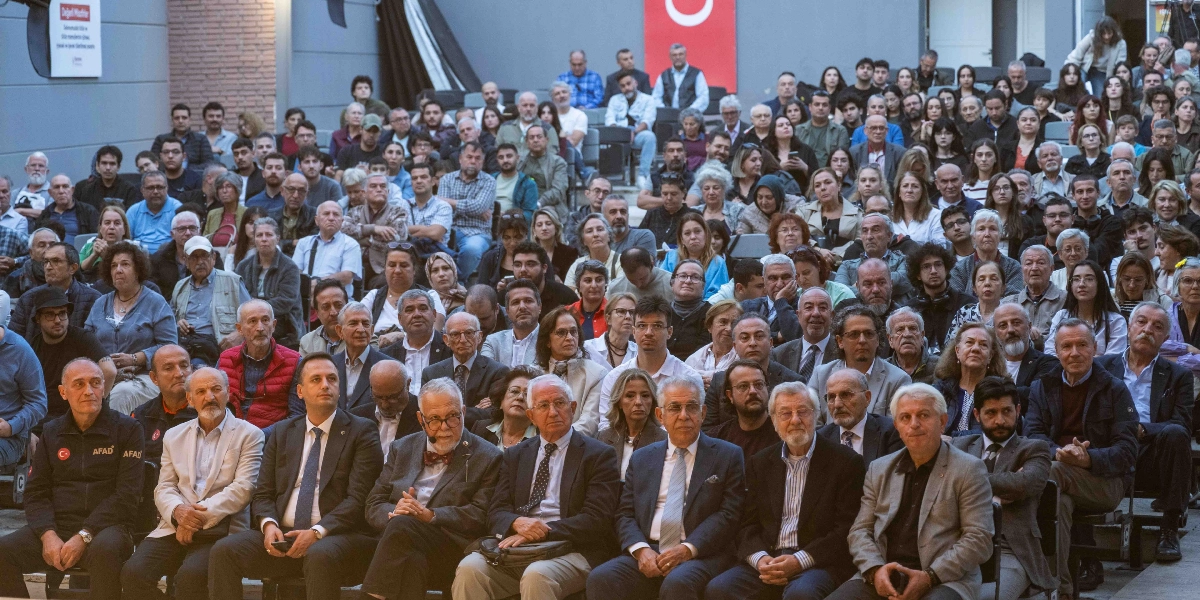 Bornova'da düzenlenen bir etkinlikte, amfi düzenindeki koltuklarda oturan kalabalık bir izleyici grubu. Ön sırada takım elbiseli protokol üyeleri ve AFAD logolu kıyafet giymiş bir kişi yer alırken, arkadaki sıralarda farklı yaş gruplarından çok sayıda katılımcı dikkatle sahneye doğru bakıyor. Salonun arka duvarında asılı Türk bayrağı ve sol üst köşede Bornova Belediyesi'ne ait bilgilendirme panosu bulunuyor.