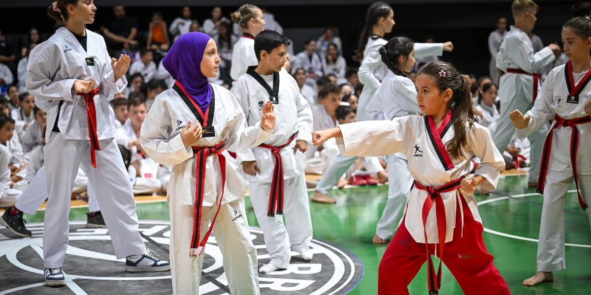 Kapalı bir spor salonunda, beyaz tekvando kıyafetleri ve kırmızı kuşakları giymiş çocuklar ve gençler bir antrenman sırasında duruş sergiliyor. Ön planda, mor başörtülü bir kız çocuğu savunma duruşunda beklerken, hemen yanındaki bir diğer kız çocuğu ileriye doğru yumruk atma hareketi yapıyor. Arka planda ise benzer kıyafetlerle antrenmana katılan çok sayıda sporcu ve onları izleyen kalabalık bir grup yer alıyor.