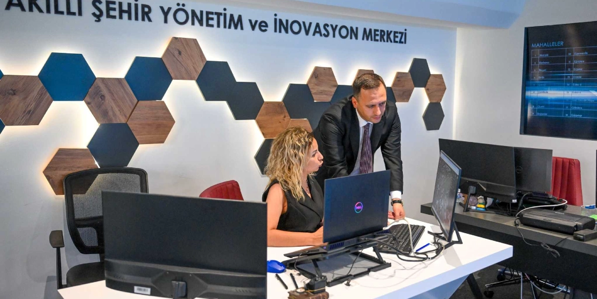 Akıllı Şehir Yönetim ve İnovasyon Merkezi yazılı bir duvarın önünde, beyaz bir çalışma masasında oturan sarı saçlı bir kadın ve onun yanında ayakta durarak bilgisayar ekranını inceleyen takım elbiseli bir erkek çalışıyor. Arka planda altıgen şeklinde ahşap ve koyu renkli dekoratif paneller, sağ tarafta ise mahalle isimlerinin listelendiği dijital bir ekran bulunuyor.