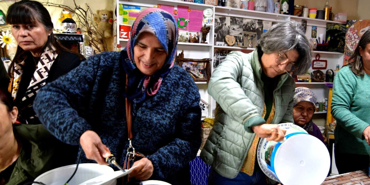 İç mekanda bir atölye çalışmasında bir araya gelen kadınlar, plastik kovalara delik açarak kompost kovası hazırlıyor. Başörtülü bir kadın elindeki havya ile kovanın kapağını delerken, yanında gri saçlı ve gözlüklü bir kadın da benzer bir plastik kovayı tutuyor. Arka planda raflarda çeşitli dekoratif objeler, fotoğraflar ve oyuncaklar yer alıyor.