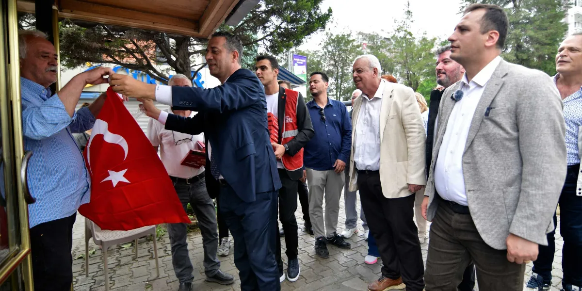 Koyu renk takım elbiseli bir adam, bir binanın girişinde elinde Türk bayrağı tutan yaşlı bir adama yardım ederken, çevresinde beyaz ceketli, mavi gömlekli ve kırmızı yelekli kişilerden oluşan bir grup izleyici yer alıyor.