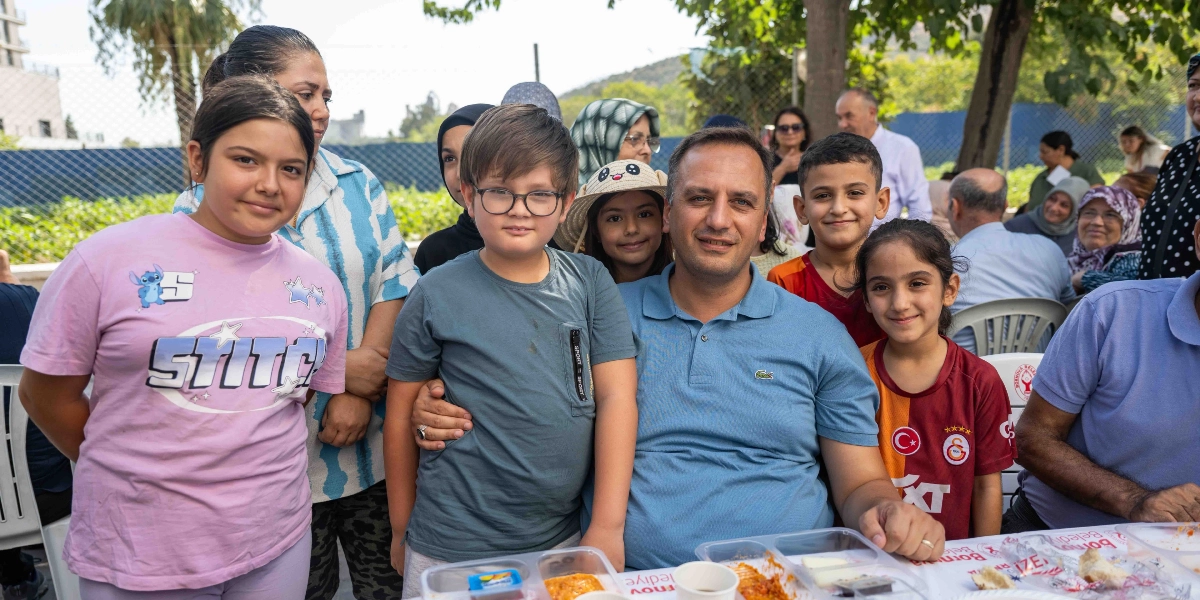 Altındağ Kent Bostanı'ndaki etkinlikte, masanın etrafında bir araya gelmiş çocuklar ve yetişkinler gülümseyerek poz veriyor. Ön planda, üzerinde 
