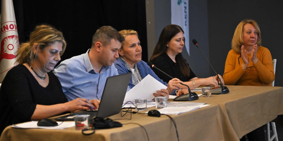Bej renkli bir masa etrafında yan yana oturan beş kişilik bir grup, bir toplantı veya panelde yer alıyor. En solda siyah kıyafetli bir kadın dizüstü bilgisayarda çalışırken, yanında açık mavi gömlekli bir erkek, onun yanında mavi ceketli bir kadın konuşma yapıyor. Yanlarında siyah kıyafetli bir başka kadın ve en sağda sarı kazaklı, elleriyle alkışlayan bir kadın bulunuyor. Masanın üzerinde mikrofonlar, belgeler ve bardaklar yer alıyor. Arka planda Bornova Belediyesi'ne ait olduğu anlaşılan logolu bir bayrak ve kısmen görünen bir panel afişi bulunuyor.