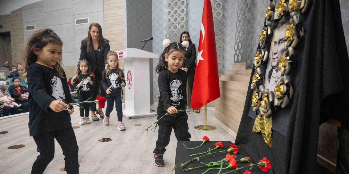 Üzerinde Atatürk baskılı siyah tişörtler giyen küçük çocuklar, bir anma töreninde ellerindeki kırmızı karanfilleri Atatürk portresinin önüne bırakıyor. Arka planda Türk bayrağı ve Bornova Belediyesi kürsüsü yer alırken, çocuklar sırayla ilerleyerek portrenin önündeki alana çiçeklerini yerleştiriyor.