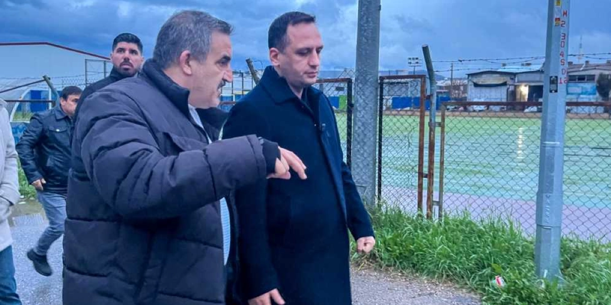 Koyu renkli kışlık mont giymiş iki erkek, açık havada, tel örgülerle çevrili yeşil bir spor sahasının yanında yürürken bir şeyler konuşuyor. Öndeki kişi eliyle sahayı işaret ederken, yanındaki kişi onu dinliyor. Arka planda ise siyah montlu başka bir kişi daha görülüyor.
