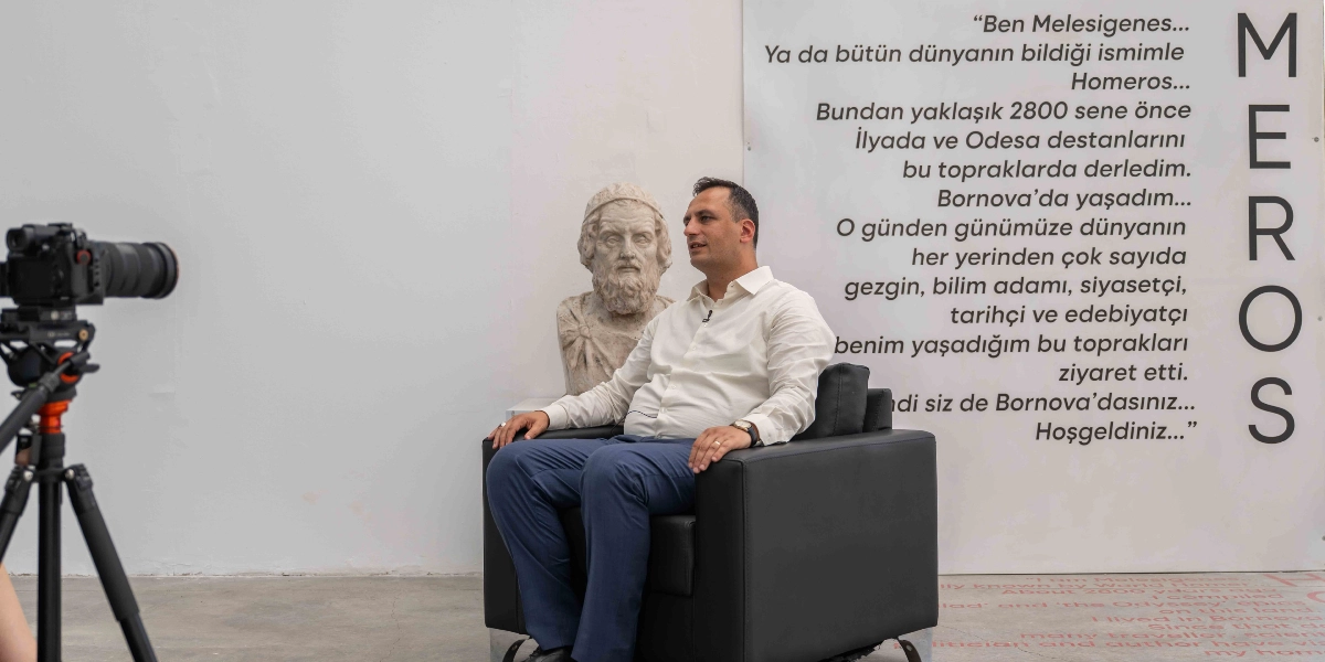 Beyaz bir duvarın önünde, siyah deri bir koltukta oturan beyaz gömlekli ve lacivert pantolonlu bir erkek, bir kamera karşısında konuşma yapıyor. Adamın hemen arkasında, Homeros'a ait olduğu düşünülen mermer bir büst yer alıyor. Duvarın sağ tarafında dikey olarak 