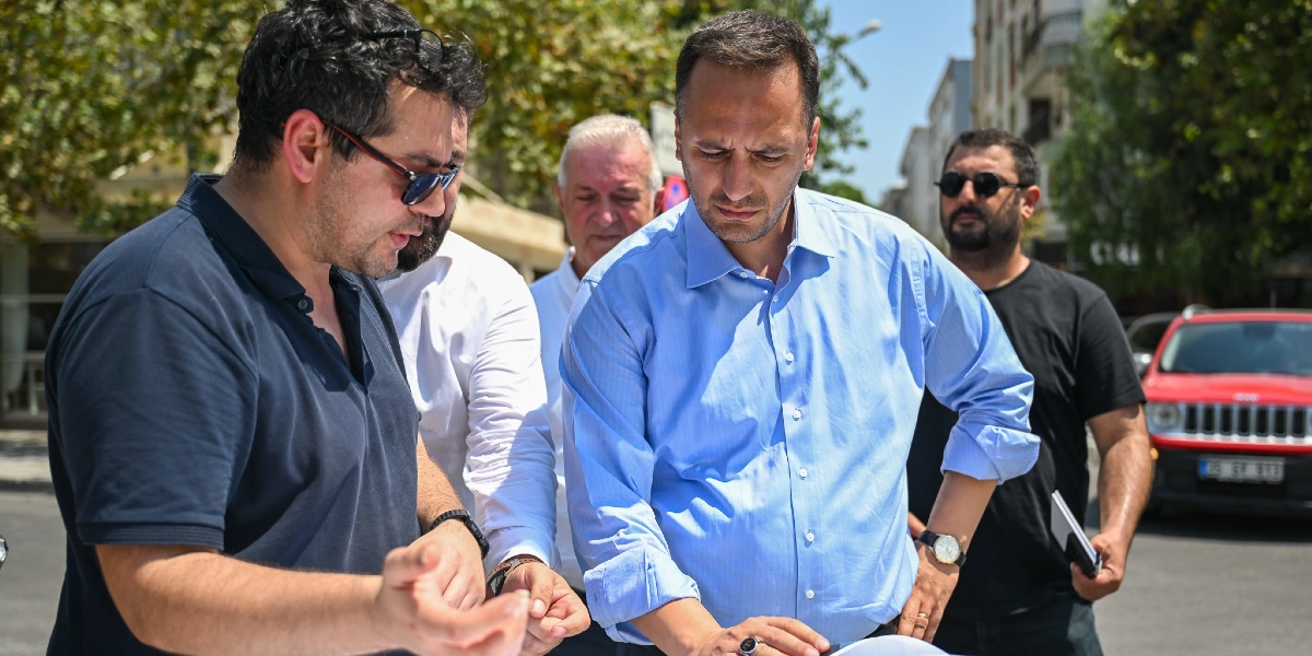 Açık havada, Bornova merkezinde bir proje üzerinde incelemelerde bulunan dört kişilik bir grup. Ön planda, üzerinde güneş gözlüğü olan koyu renk tişörtlü bir adam, yanında duran ve açık mavi gömlek giyen bir yetkiliye bir şeyler anlatıyor. Grubun geri kalanında beyaz gömlekli bir kişi ve siyah tişörtlü bir başka erkek daha yer alıyor. Arka planda ise ağaçlar ve park halindeki kırmızı bir araç görülüyor.