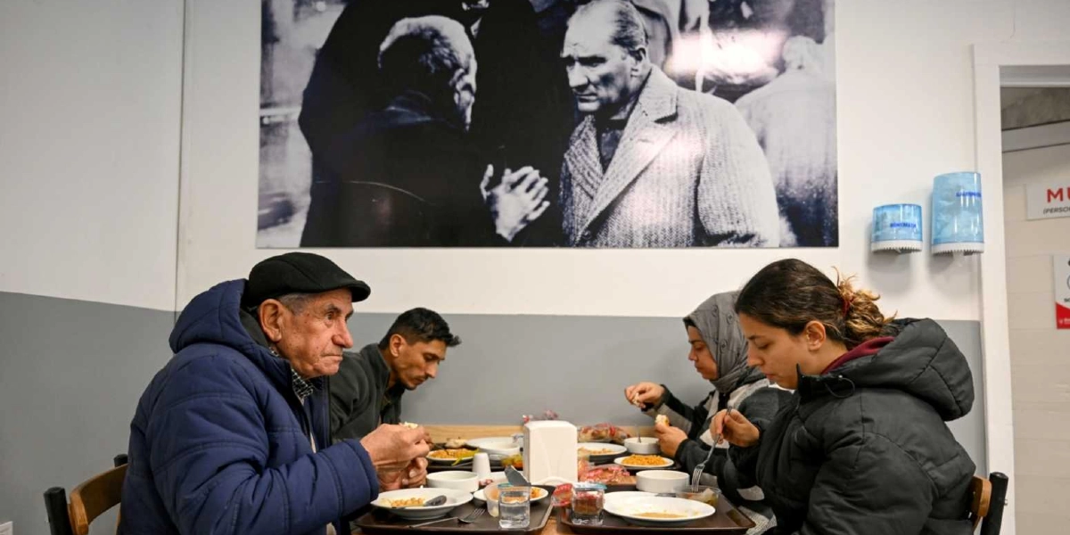 İç mekanda bir masada yemek yiyen dört kişilik bir grup görülüyor. Ön planda kasketli yaşlı bir adam, yanında genç bir erkek, karşısında ise başörtülü bir kadın ve genç bir kadın oturuyor. Masada tabaklar, bardaklar ve peçetelik bulunuyor. Arka plandaki duvarda, Mustafa Kemal Atatürk'ün bir vatandaşla sohbet ederken çekilmiş büyük, siyah beyaz bir fotoğrafı asılı duruyor.
