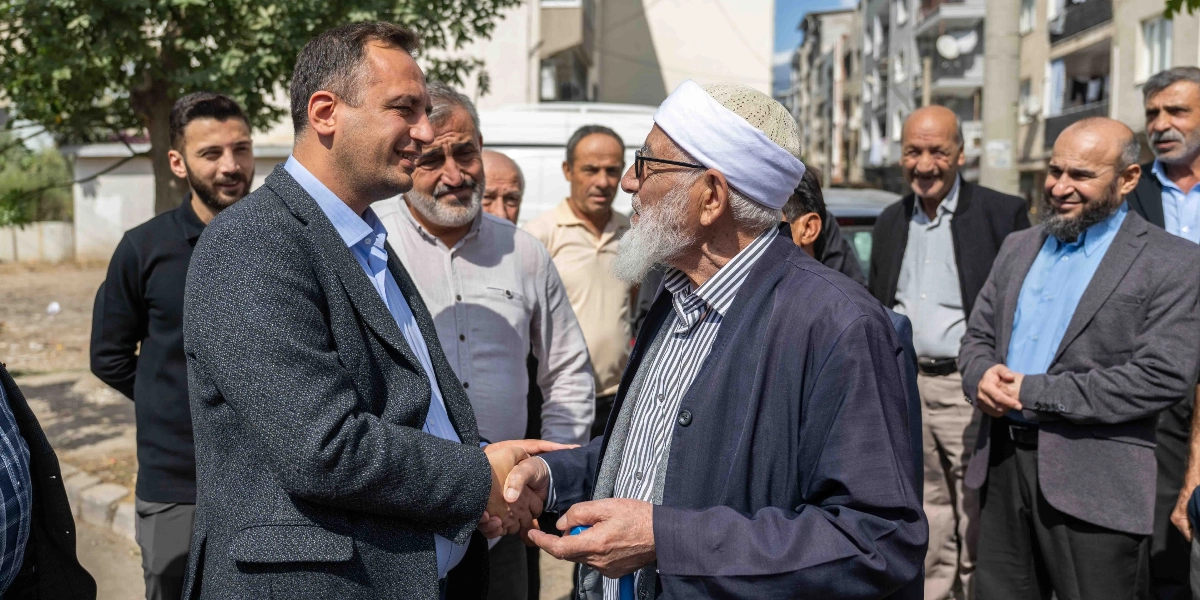 Koyu renk ceketli ve açık mavi gömlekli bir erkek, sokakta beyaz takkeli, beyaz sakallı ve gözlüklü yaşlı bir adamla el sıkışıyor. İkilinin çevresinde onları gülümseyerek izleyen bir grup erkek bulunuyor. Arka planda ise binaların olduğu bir mahalle sokağı yer alıyor.