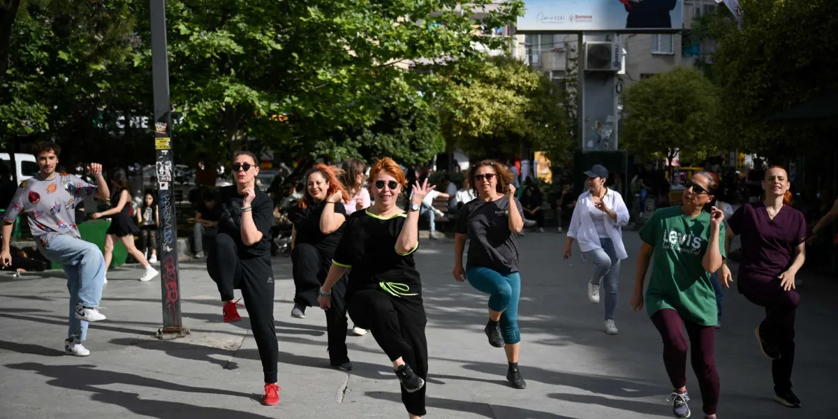 Bornova'da güneşli bir günde, ağaçlıklı bir meydanda bir araya gelmiş sekiz kişilik bir grup, müzik eşliğinde senkronize bir şekilde dans ediyor. Farklı yaş ve tarzlarda giyinmiş katılımcılar, neşeli bir ifadeyle aynı koreografiyi uygularken, arka planda günlük yaşamın devam ettiği sokak ve ağaçlar görülüyor.