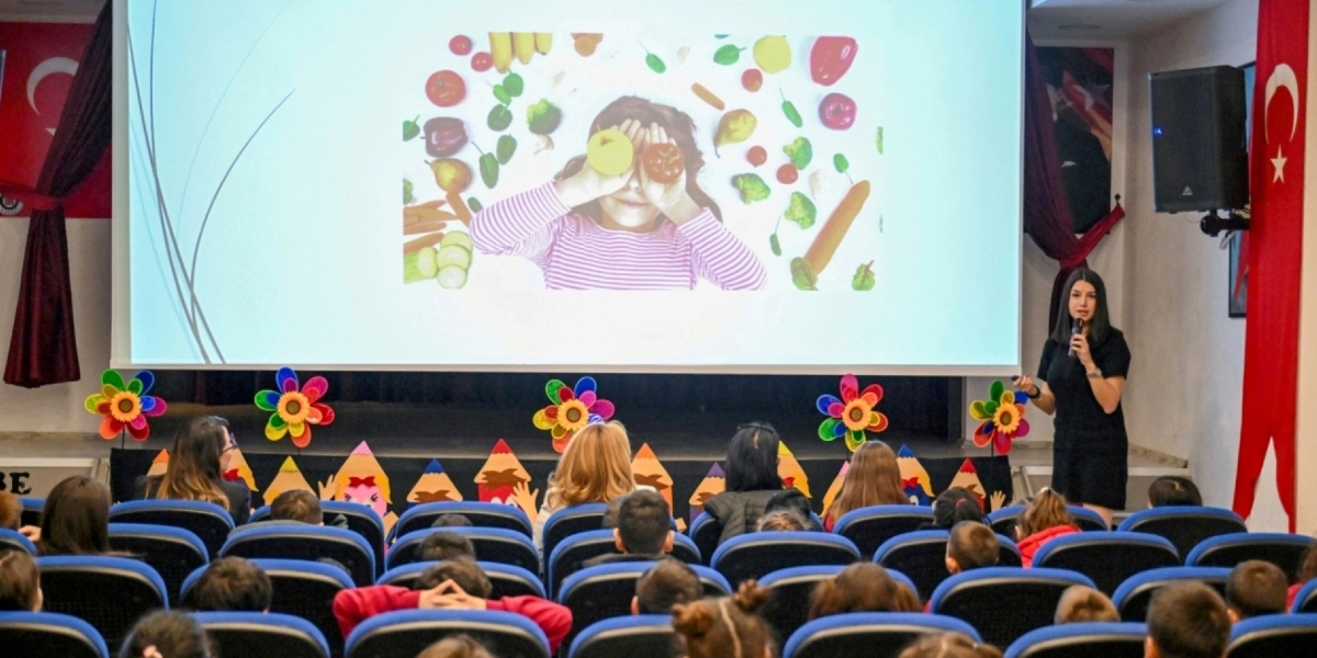 Konferans salonunda, sahnedeki büyük ekranda gözlerinin önünde bir elma ve bir portakal tutan bir çocuk ile etrafında çeşitli sebze ve meyvelerin olduğu bir görsel yansıtılırken, siyah kıyafetli bir kadın mikrofonla öğrencilere sunum yapıyor. Salonun ön kısmında renkli çiçek figürleri ve kalem şeklinde süslemeler yer alırken, öğrenciler mavi koltuklarda oturarak sunumu izliyor. Salonun iki yanında Türk bayrakları asılı bulunuyor.