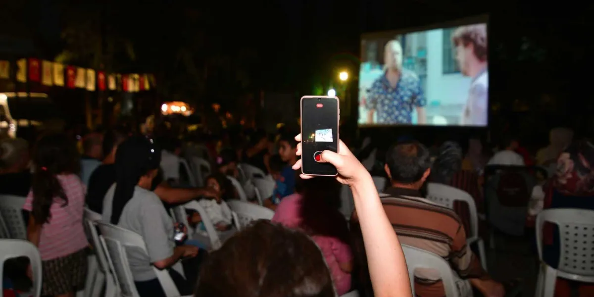 Gece vakti açık havada kurulan bir sinema ekranında film izleyen kalabalık bir grup. İzleyicilerin önünde oturan bir kişi, elindeki akıllı telefonla dev ekrandaki film sahnesini kaydediyor. Ekranın arka planında beyaz plastik sandalyelerde oturan izleyiciler ve yukarıda asılı duran süs bayrakları görülüyor.
