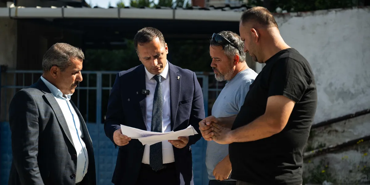 Dış mekanda, takım elbiseli ve kravatlı bir yetkilinin elindeki belgeleri incelediği, yanında bulunan iki kişiyle birlikte bir proje veya çalışma üzerine görüştüğü an.