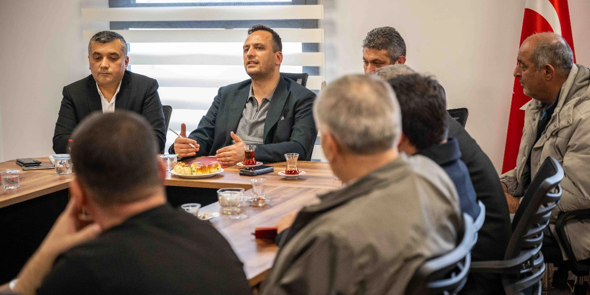 Başkan Ömer Eşki, bir toplantı masasında karşısında oturan muhtarlara hitap ediyor. Masanın başında oturan Eşki, ellerini kullanarak konuşurken, solunda takım elbiseli bir kişi, sağında ise diğer katılımcılar yer alıyor. Masanın üzerinde çay bardakları ve küçük bir tabak içerisinde atıştırmalıklar bulunuyor. Arka planda ise bir Türk bayrağı yer alıyor.