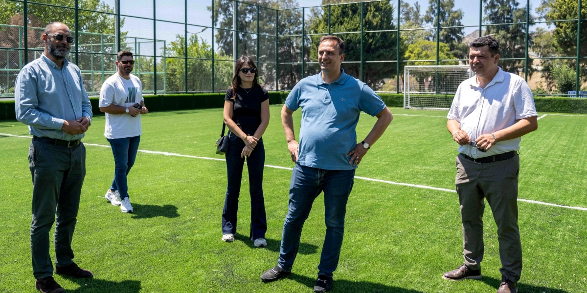 Açık havada, güneşli bir günde suni çim kaplı bir futbol sahasında duran beş kişilik bir grup. Gruptaki üç erkek ve bir kadın günlük kıyafetler içinde, bir erkek ise beyaz tişört ve kot pantolonla sahada yürürken görülüyor. Arka planda tel örgülerle çevrili saha ve ağaçlar yer alıyor.