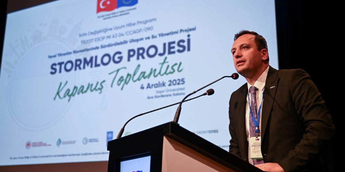 Koyu renk takım elbiseli, kravatlı bir konuşmacı, kürsüde mikrofon başında hitap ediyor. Arkasındaki dev ekranda, üzerinde Türkiye Cumhuriyeti ve Avrupa Birliği bayraklarının yer aldığı, 