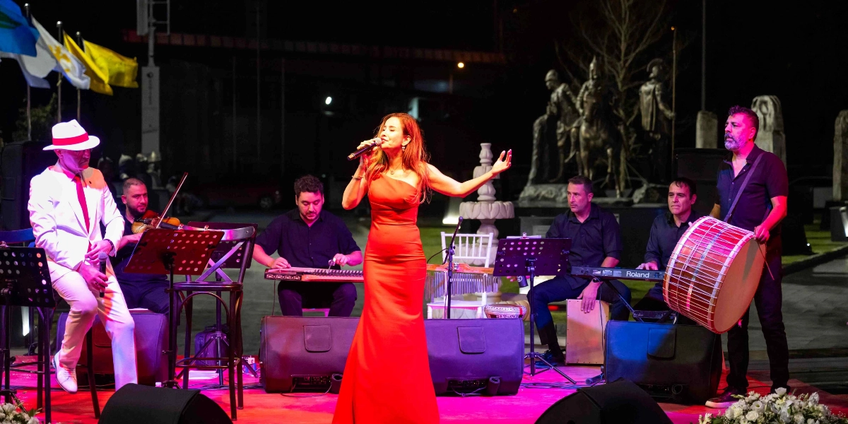 Gece vakti açık havada düzenlenen bir etkinlikte, kırmızı uzun bir elbise giymiş kadın solist mikrofonla şarkı söylerken, ona eşlik eden müzisyenler sahne alıyor. Müzisyenlerden biri beyaz takım elbise ve şapka ile oturarak eşlik ederken, diğerleri keman, kanun, klavye ve davul çalıyor. Sahnenin arka planında aydınlatılmış bir heykel grubu ve bayraklar yer alıyor.
