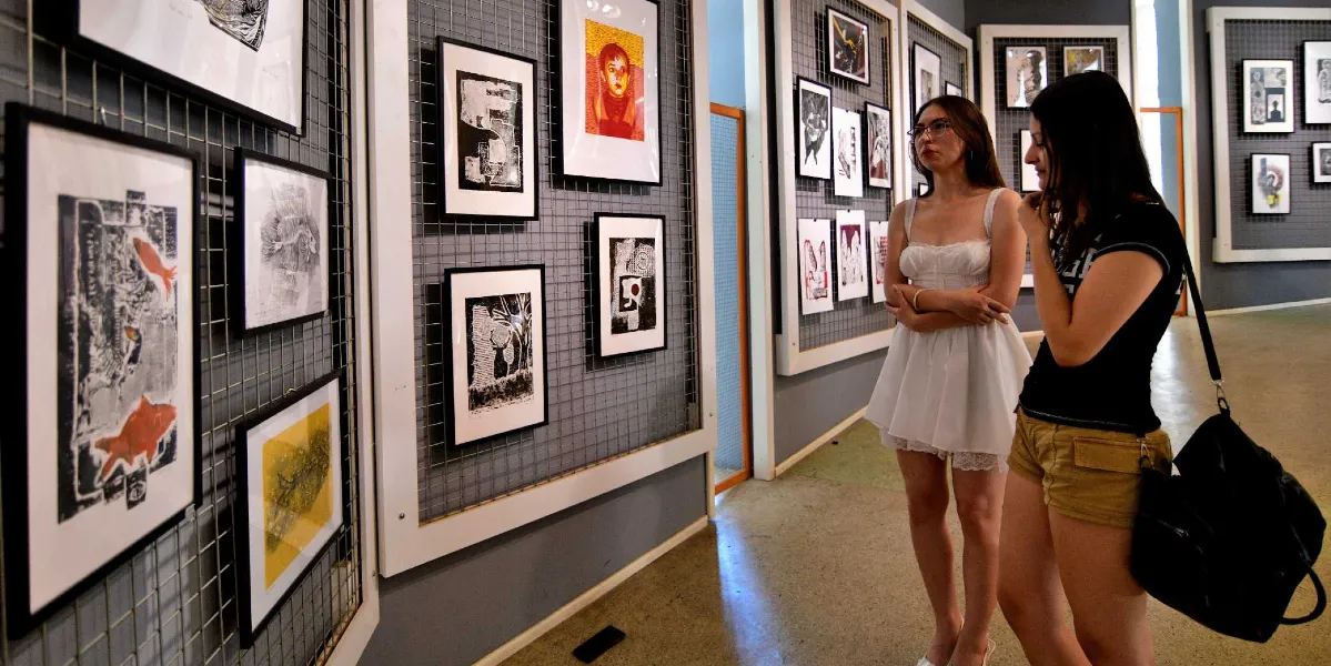 Bir sanat galerisinde, ızgara panellere asılmış çeşitli siyah beyaz ve renkli baskı eserleri inceleyen iki genç kadın. Beyaz elbiseli kadın kollarını kavuşturmuş eserlere bakarken, yanındaki siyah tişörtlü ve omuz çantalı kadın ona doğru dönmüş bir şeyler anlatıyor.