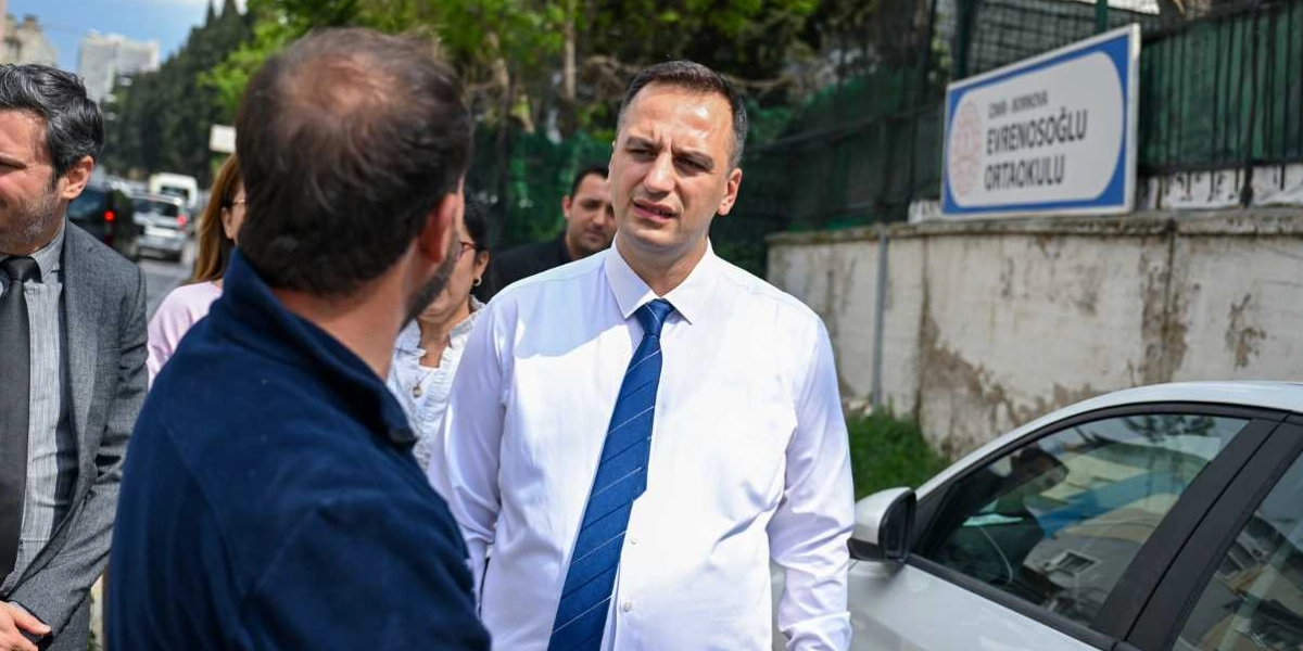Beyaz gömlekli ve mavi kravatlı bir erkek, Bornova Evrenosoğlu Ortaokulu tabelasının önünde bir grupla sohbet ediyor.